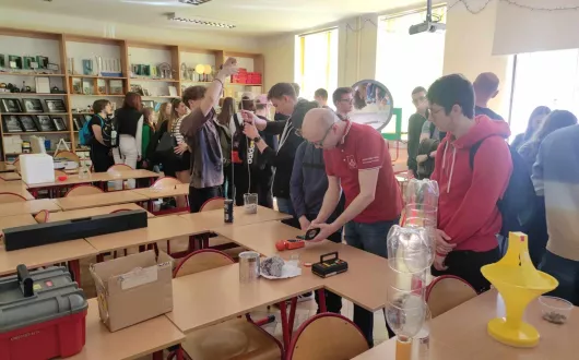 Zdjęcie przedstawia doktora inżyniera Piotra Słomę wraz z uczniami liceum w pracowni fizycznej podczas wykonywania doświadczenia. 