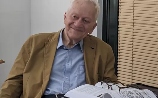 Starszy, uśmiechnięty mężczyzna ubrany w koszulę i marynarkę siedzi przy biurku, na którym leżą otwarte książki i okulary. 