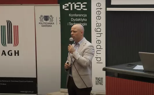 Mężczyzna w jasnej marynarce stoi przed publicznością i mówi do mikrofonu podczas konferencji dydaktycznej organizowanej przez AGH i Politechnikę Gdańską. W tle widoczne są banery z logotypami uczelni oraz adresem strony etee.agh.edu.pl.