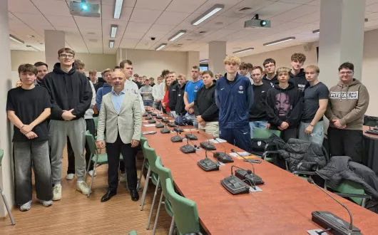 Grupa kilkudziesięciu chłopców stoi w sali konferencyjnej przy długim stole z mikrofonami. Na pierwszym planie, pośrodku, stoi mężczyzna w jasnej marynarce i koszuli. 