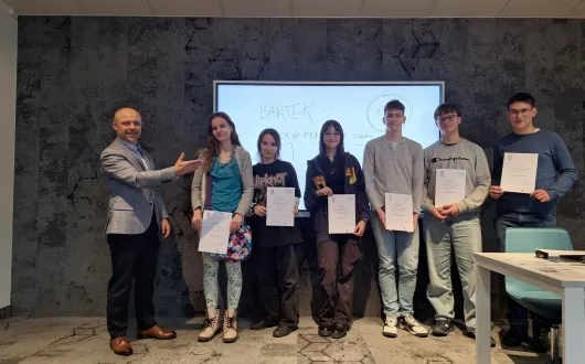 Zdjęcie grupowe w sali wykładowej przedstawiające kilka osób stojących w rzędzie i trzymających certyfikaty. Obok nich stoi prowadzący, który gestem wskazuje na uczestników. W tle widoczna jest ekran.