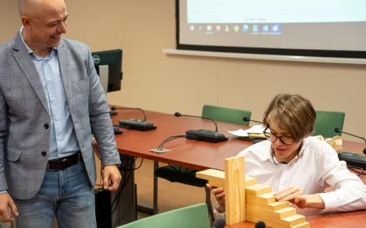 W sali wykładowej prowadzący zajęcia obserwuje ucznia podczas układania drewnianej konstrukcji schodkowej na stole. W tle widoczne są stoły konferencyjne, mikrofony i ekran projekcyjny.