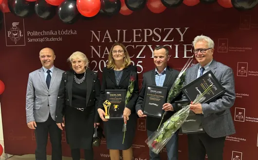 Grupowe zdjęcie kilku osób stojących przed ścianką z napisem „Najlepszy Nauczyciel”. Część osób trzyma dyplomy i kwiaty. Nad nimi widoczna jest dekoracja z czerwono-czarnych balonów.
