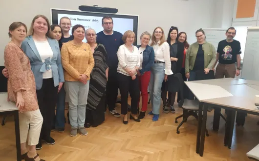 Grupa osób stoi w sali szkoleniowej. Za nimi widać duży ekran, tablicę i stoły. Uczestnicy pozują do wspólnego zdjęcia.