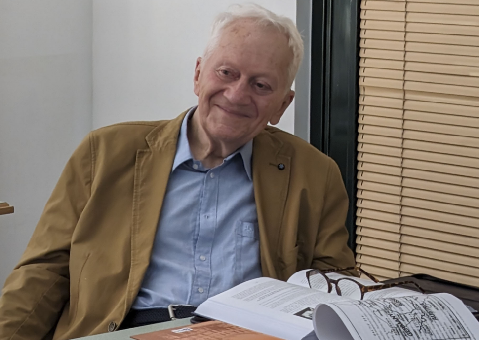 Starszy, uśmiechnięty mężczyzna ubrany w koszulę i marynarkę siedzi przy biurku, na którym leżą otwarte książki i okulary. 