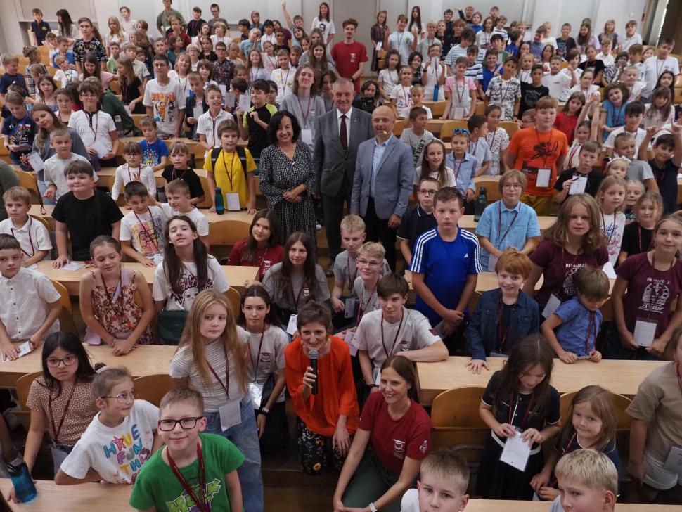 Duża grupa dzieci i kilku dorosłych pozuje do wspólnego zdjęcia w sali wykładowej. Dzieci siedzą i stoją między ławkami, uśmiechają się w stronę aparatu. W pierwszym rzędzie kilka osób dorosłych, w tym kobieta z mikrofonem.
