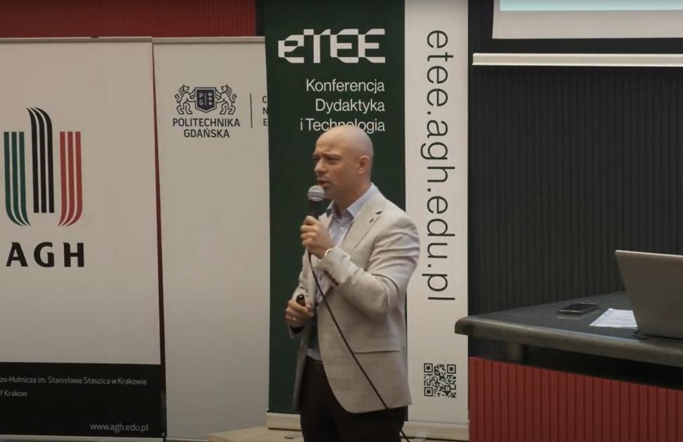 Mężczyzna w jasnej marynarce stoi przed publicznością i mówi do mikrofonu podczas konferencji dydaktycznej organizowanej przez AGH i Politechnikę Gdańską. W tle widoczne są banery z logotypami uczelni oraz adresem strony etee.agh.edu.pl.