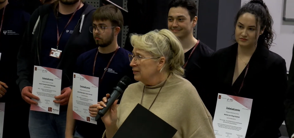 Kobieta mówi do mikrofonu podczas konferencji. Wokół niej stoją studenci, którzy trzymają dyplomy.