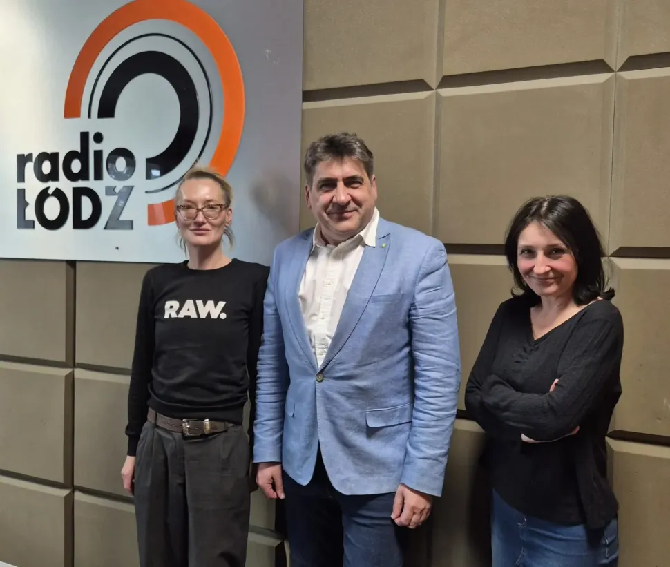 Trzy osoby stoją w studiu radiowym przy ścianie z logo Radia Łódź. W środku stoi mężczyzna w jasnoniebieskiej marynarce, a po jego bokach stoją dwie kobiety w ciemnych bluzach.