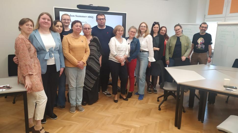 Grupa osób stoi w sali szkoleniowej. Za nimi widać duży ekran, tablicę i stoły. Uczestnicy pozują do wspólnego zdjęcia.