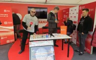 Osoba z mikrofonrm trzyma mikrofon, stojąc przy stole na którym stoją puszki. Całość umieszczona jest pod namiotem wystawowym graficznie oznaczonym Polskie Radio