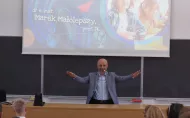 Mężczyzna w jasnej marynarce stoi przed tablicą w sali wykładowej i gestykuluje z rozłożonymi rękami, mówiąc do uczniów. Za nim widoczny jest ekran z prezentacją zatytułowaną „MateMagia?”, z imieniem i nazwiskiem dr. Marek Małolepszy, prof. PŁ oraz datą 20.09.2025.