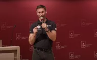 Mężczyzna w granatowej koszulce z logo ESA stoi na tle bordowej ściany z napisem Politechnika Łódzka. Trzyma mikrofon i gestykuluje podczas wystąpienia.