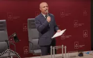 Mężczyzna w garniturze stoi na tle bordowej ściany z logo Politechniki Łódzkiej. Trzyma mikrofon i kartki w ręku, przemawia podczas wydarzenia. Przed nim na stole znajdują się rekwizyty do doświadczeń fizycznych.